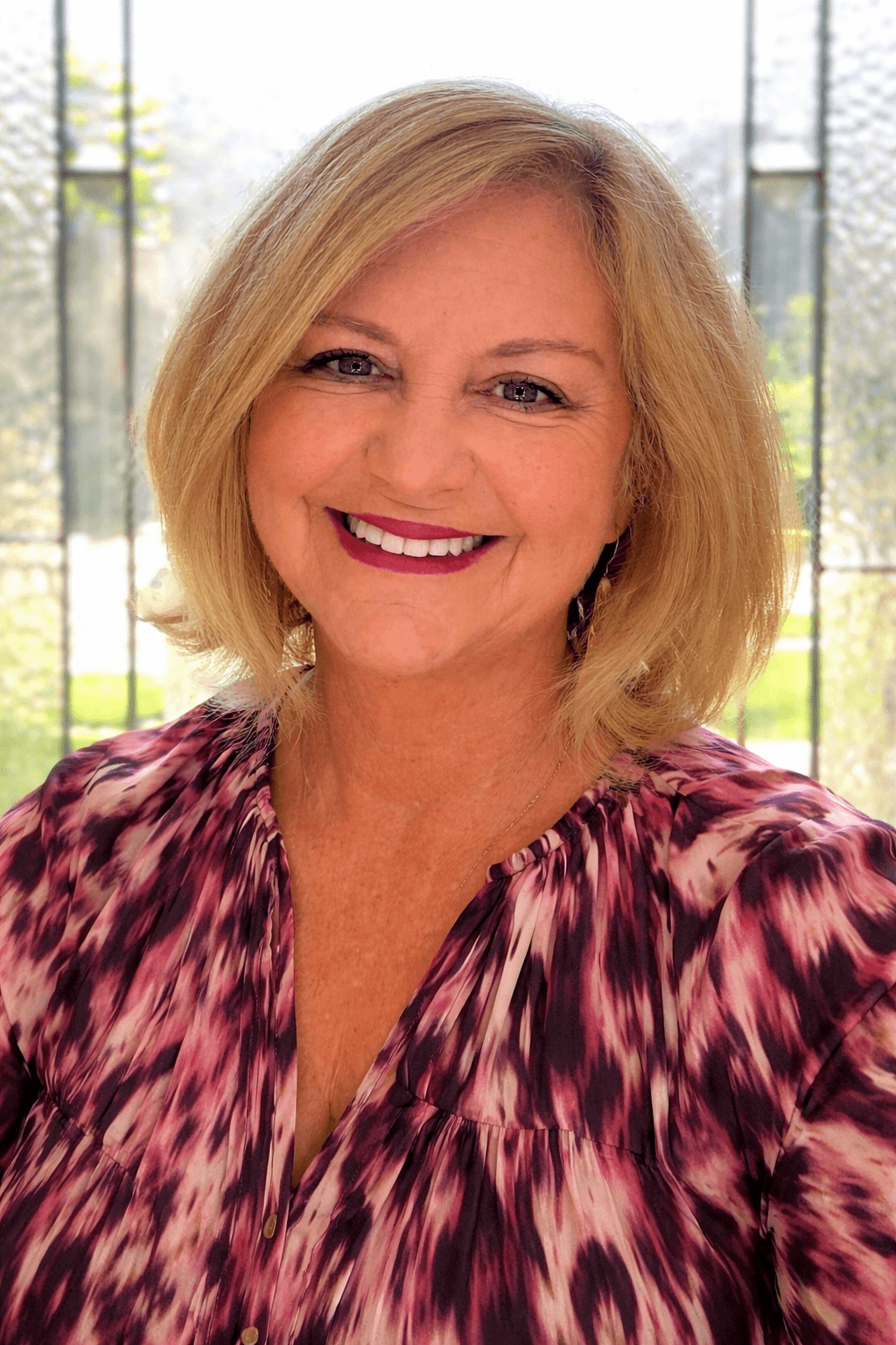 Smiling blonde woman with shoulder-length hair wearing a purple and pink abstract patterned blouse.